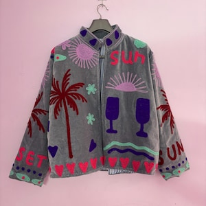 Puede incluir: Chaqueta gris con bordados de palmeras, corazones y las palabras "SUN" y "SET" en rosa. La chaqueta tiene un cuello alto y un forro a rayas, con detalles morados y rosas.