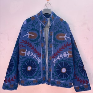 May include: A blue embroidered jacket with floral and leaf patterns in shades of brown, red, and green. The jacket is open, revealing a striped lining. It hangs on a hanger against a pink wall.