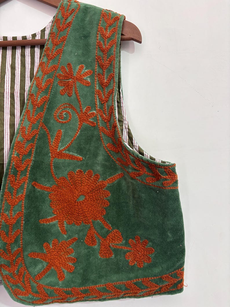 Puede incluir: Un chaleco de terciopelo verde con intrincados bordados florales en naranja. El chaleco presenta un forro a rayas y se exhibe en una percha de madera. El bordado incluye patrones florales y de hojas, agregando un toque decorativo.
