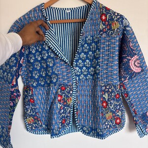 May include: A blue quilted jacket with floral and geometric patterns. The jacket features a V-neck and is lined with blue and white striped fabric. Red and pink floral accents are visible throughout the design.