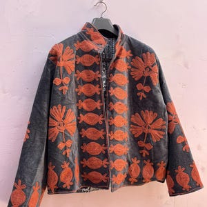 May include: A dark gray jacket with orange floral embroidery. The jacket has a high collar and a zipper closure. The embroidery features stylized flowers and other decorative elements. The jacket is hanging on a black hanger.