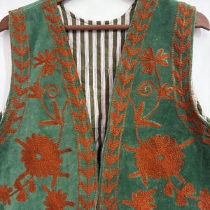 Puede incluir: Un chaleco verde con bordados florales y ribetes de color &oacute;xido. El forro interior del chaleco presenta un estampado a rayas en verde, blanco y rojo. El chaleco est&aacute; colgado de una percha de madera.