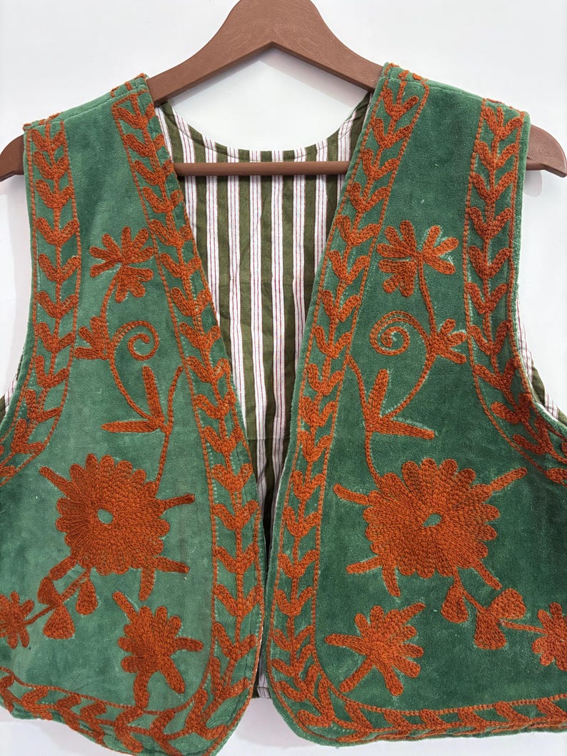 Puede incluir: Un chaleco verde con bordados florales y ribetes de color &oacute;xido. El forro interior del chaleco presenta un estampado a rayas en verde, blanco y rojo. El chaleco est&aacute; colgado de una percha de madera.