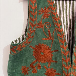 Puede incluir: Un chaleco verde con bordado floral color &oacute;xido. El chaleco tiene un forro a rayas en tonos verdes y blancos. El bordado presenta patrones detallados de flores y hojas, agregando un toque decorativo a la prenda.