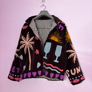 Puede incluir: Chaqueta marrón con diseños bordados coloridos de palmeras, corazones y la palabra "SUN". La chaqueta tiene un forro a rayas y está colgada de una percha gris.