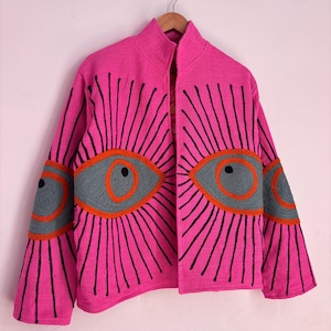 May include: A vibrant pink jacket with a high collar and open front. The jacket features embroidered eye designs in gray, orange, and black on the sleeves and chest. The jacket is hanging on a wooden hanger against a light pink background.