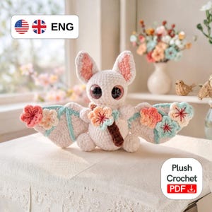 May include: A white crochet bat plushie with large, expressive eyes and pink-lined ears. The bat's wings are adorned with pastel-colored crochet flowers and a brown stem. The plushie is a handmade item, perfect for a nursery or as a unique gift.