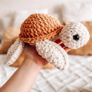 Puede incluir: Una tortuga de crochet hecha a mano. La tortuga tiene un caparazón texturizado de color marrón anaranjado, una cabeza blanca y aletas blancas. El juguete tiene un ojo negro y está hecho de hilo suave. Un artículo lindo y tierno.