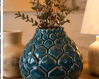 May include: A round, teal-colored ceramic vase with a textured, honeycomb-like pattern across its surface. The vase has a glossy finish with darker shading in the recessed areas between the raised, hexagonal shapes and holds dried plant stems.