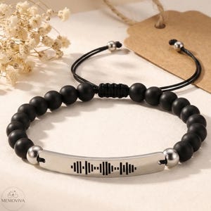 Puede incluir: Pulsera de cuentas negras con una barra plateada con un diseño de onda de sonido. La pulsera tiene un cierre de nudo corredizo y cuentas plateadas. Un accesorio elegante.