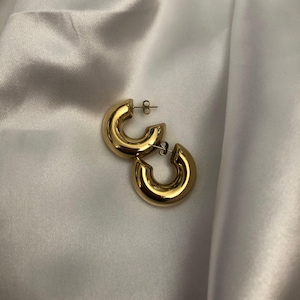 Pode incluir: Um par de brincos de argola dourados e grossos. Os brincos têm um design polido e arredondado, refletindo a luz. Os brincos são exibidos em um tecido branco acetinado.