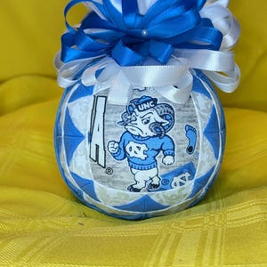 May include: A spherical fabric ornament featuring a quilted pattern of light blue and white fabric panels. The center panel displays a graphic of a ram mascot wearing a blue sweater with the letters UNC. A large bow of blue and white ribbons sits on top.