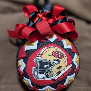 May include: A spherical fabric ornament featuring a San Francisco 49ers football helmet graphic on a red circular patch. The ornament is constructed with folded fabric segments in black, red, and gold, topped with a decorative red and black ribbon bow.