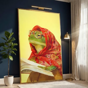 May include: A framed art print featuring a green frog wearing glasses and a red floral scarf, reading a book. The frog is set against a yellow background, with a gold frame. The artwork is displayed in a room with a plant and a floor lamp.