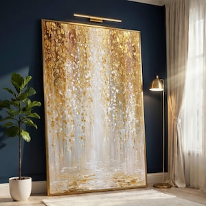 Op de afbeelding: Groot abstract schilderij met een gouden lijst. Het kunstwerk toont een waterval van gouden en witte penseelstreken, waardoor een textuureffect ontstaat. Het schilderij wordt tentoongesteld in een kamer met een plant en een vloerlamp.