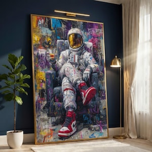 May include: Large framed artwork featuring an astronaut in a spacesuit seated in a chair, holding a drink. The astronaut wears red and white sneakers. The background is a colorful abstract design with graffiti-style elements. The artwork has a gold frame.