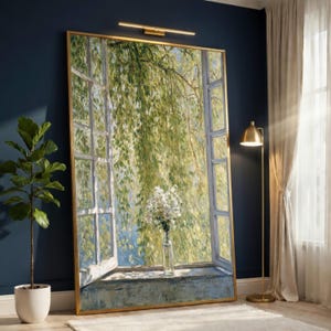 May include: Large framed artwork depicting a window scene with a view of lush green foliage. A vase of white flowers sits on the windowsill. The artwork has a gold frame and is displayed in a room with a blue wall and a potted plant.