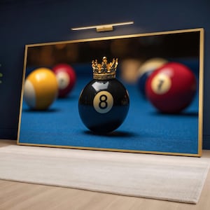 May include: A framed print showing a black eight ball with a gold crown, set on a blue pool table. Other billiard balls are in the background. The frame is gold.