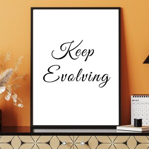 May include: A framed print with the words "Keep Evolving" in elegant script. The black frame surrounds a white background. A black desk lamp, a calendar, and a decorative vase with dried flowers are also visible.