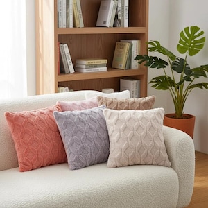 May include: A collection of decorative throw pillows in various colors, including coral, lavender, and beige. The pillows have a textured, geometric pattern and are arranged on a white sofa. A wooden bookshelf and potted plant are in the background.