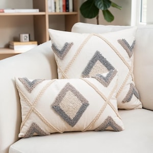 Könnte beinhalten: Zwei dekorative Kissen mit cremefarbenem Hintergrund. Die Kissen haben ein Rautenmuster mit grau-beigen strukturierten Akzenten und geflochtenen Zierleisten. Die Kissen liegen auf einem weißen Sofa.