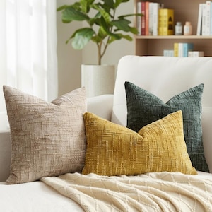 May include: Four textured throw cushions in neutral shades are arranged on a white sofa. The cushions include beige, mustard yellow, and dark teal green. The woven texture adds a touch of elegance to the living space, creating a cozy atmosphere.