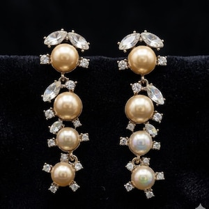 May include: A pair of gold-toned drop earrings. Each earring features a large, round, faux pearl at the top, followed by three smaller pearls. Clear, sparkling crystals and marquise-shaped stones accent the design, creating a glamorous look.