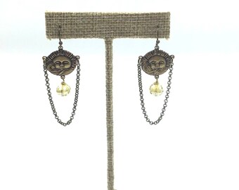 May include: A pair of gold tone earrings with a sun face charm and a dangling crystal bead. The earrings are displayed on a burlap earring stand.