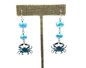 May include: A pair of dangle earrings featuring blue crystal beads and a blue crab charm. The earrings are displayed on a burlap earring stand.