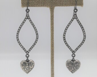May include: A pair of silver earrings with a teardrop-shaped design. The earrings are adorned with small clear stones and feature a heart-shaped charm with a floral design.