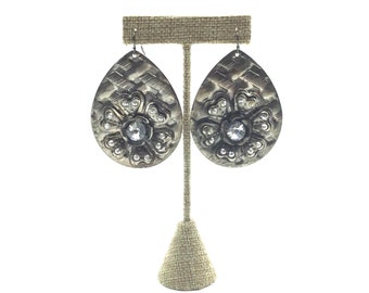 May include: A pair of silver teardrop-shaped earrings with a flower design and a clear rhinestone center. The earrings are hanging from a brown fabric stand.
