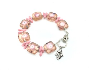 May include: A pink and clear glass bracelet with a floral design and a silver hamsa charm.