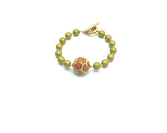 May include: A gold bracelet with a toggle clasp and a large, patterned bead. The bead is green, yellow, and orange.