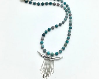 May include: A beaded necklace with turquoise and gray stones. The necklace features a silver pendant with a bull horn design and dangling silver chains. The necklace is displayed on a white background.