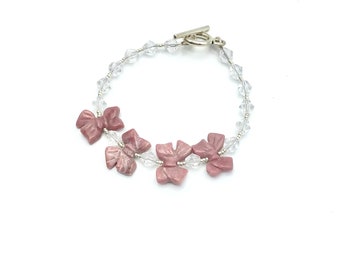 May include: A delicate bracelet featuring a silver clasp and a strand of clear crystal beads. Three pink bow-shaped charms are attached to the strand.