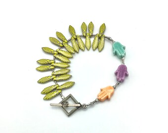 May include: A green, purple, and orange hamsa hand charm bracelet with a silver clasp and yellow patterned beads.
