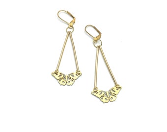 May include: A pair of gold-colored earrings. Each earring features a butterfly-shaped charm at the bottom, with a long, slender, triangular design. The earrings have a lever-back closure. The background is plain white.