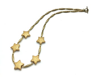May include: A gold-toned necklace featuring five textured star pendants. The stars are connected by elongated gold beads and smaller round beads. The necklace has a toggle clasp and is set against a white background.