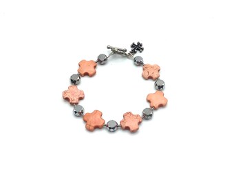 May include: A bracelet with a silver clasp and a cross charm. The bracelet is made of alternating peach-colored cross-shaped beads and silver beads.