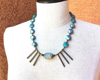 May include: A teal blue and bronze necklace with a large oval-shaped bead in the center. The necklace is made of faceted beads and has a geometric design.