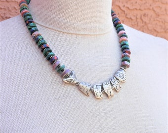 May include: A multi-colored beaded necklace with a silver fish pendant. The beads are green, pink, purple, and black. The fish pendant is made of silver and has a detailed design.