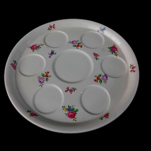 Op de afbeelding: Een witte serveerschaal met een bloemmotief. De schaal heeft een ronde vorm met zeven verzonken delen en is versierd met kleine clusters van roze, rode, paarse en blauwe bloemen. De achtergrond is zwart.