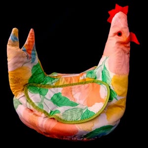 Può includere: Una teiera a forma di gallina in tessuto con un motivo floreale nei toni del pesca, giallo, verde e bianco. La gallina ha una cresta e un becco rossi e un bordo verde. La teiera è progettata per mantenere il tè caldo.