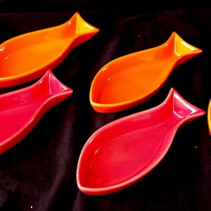 Può includere: Sei piatti da portata in ceramica a forma di pesce, nei toni del rosso e dell'arancione. I piatti hanno una finitura lucida e sono disposti su una superficie scura. Il design a forma di pesce aggiunge un tocco unico.