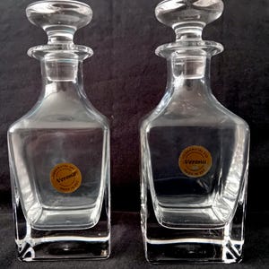 Op de afbeelding: Twee heldere glazen decanteerflessen met stoppers. Elke decanteerfles heeft een vierkante basis en een afgeronde body, met een label met de tekst "Verona" en "Made in EEC". De stoppers zijn rond en passen perfect in de halzen van de decanteerflessen.