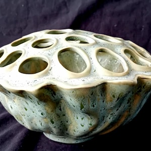 Op de afbeelding: Een keramische kom met een uniek ontwerp. De kom is lichtgroen van kleur met een textuur en een reeks ovale openingen aan de bovenkant. De bodem heeft een golvend, getextureerd patroon.