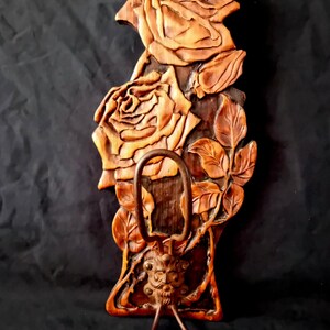 May include: Wooden wall-mounted coat rack with carved roses and leaves. The rack features a decorative metal hook and two lower hooks. The wood has a warm, natural finish, and the design is detailed and artistic.