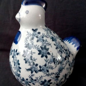 Op de afbeelding: Een keramieken kippenbeeldje met een witte basis en een blauw bloemmotief. De kip heeft een blauwe kam, lellen en staart. Het bloemmotief bedekt het lichaam van de kip en creëert een decoratief stuk.