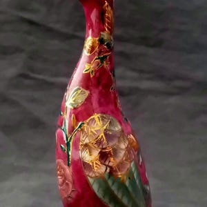 Op de afbeelding: Decoratief keramieken eendenbeeldje in een rijke bordeauxrode tint, versierd met bloemen- en bladerpatronen in goud, groen en roze. De eend heeft een lange nek en een gedetailleerde snavel, die een mix van kunst en vakmanschap laat zien.