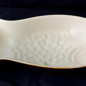 Könnte beinhalten: Eine weiße, fischförmige Servierschale aus Keramik mit einem strukturierten Schuppenmuster. Die Schale hat einen braunen Rand und eine Schwanzflosse an einem Ende. Die Schale ist etwa 30 cm lang.